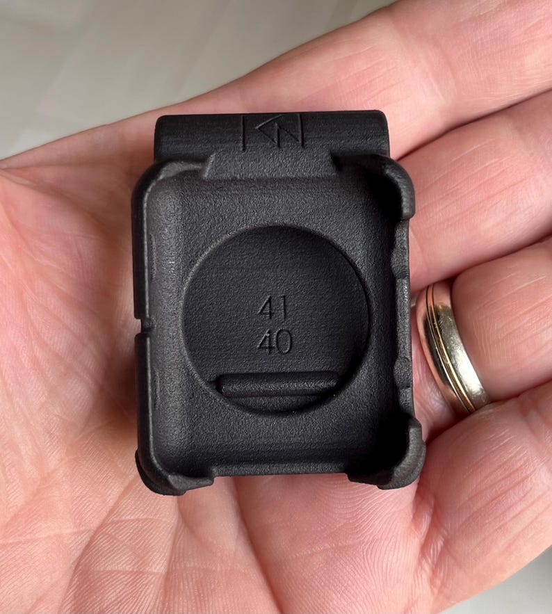 May include: A black protective case for a smartwatch, featuring a textured matte finish and a rectangular frame. The interior of the case is embossed with the numbers 41 and 40, and the top edge displays a small geometric logo symbol.