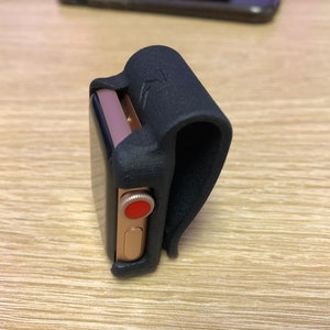 May include: A gold-toned smartwatch with a black screen and a red digital crown, secured within a black, textured, U-shaped clip-on holder. The holder features a small embossed logo on its curved exterior surface, designed to attach the watch.