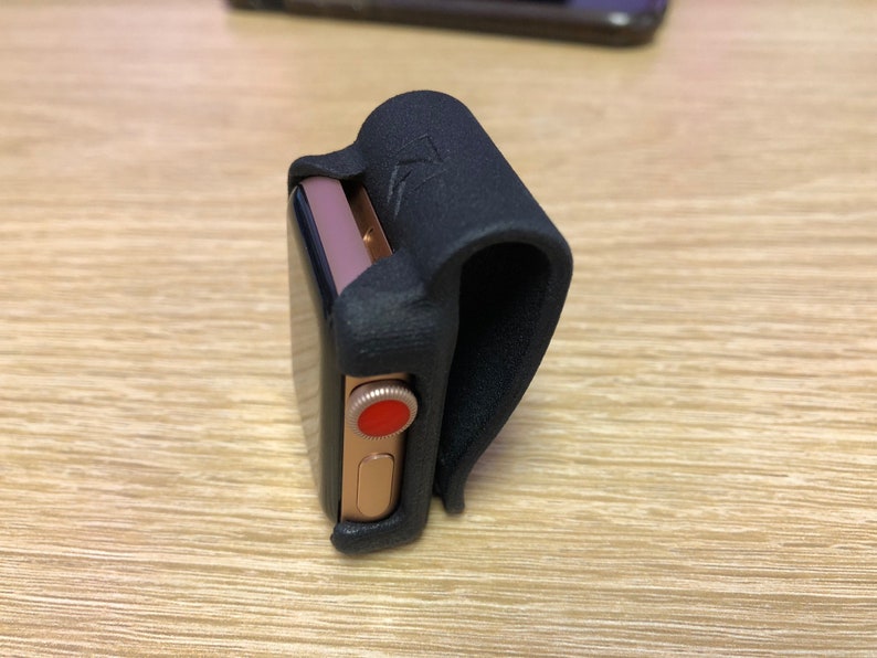 May include: A gold-toned smartwatch with a black screen and a red digital crown, secured within a black, textured, U-shaped clip-on holder. The holder features a small embossed logo on its curved exterior surface, designed to attach the watch.