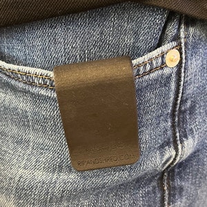 May include: A matte black rectangular money clip attached to the edge of a blue denim pocket. The clip features the embossed text "PATENT PENDING" and "RIPANDSHRED.COM" near the bottom edge, providing a secure way to carry currency or cards.