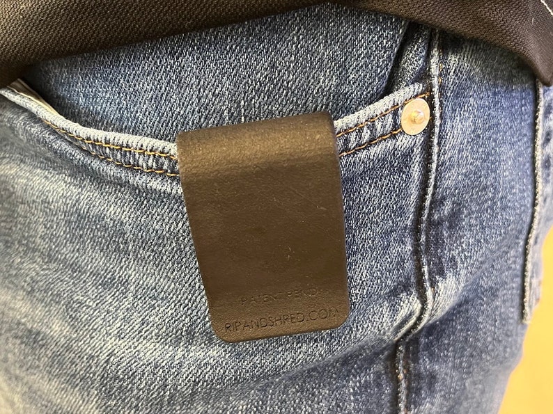 May include: A matte black rectangular money clip attached to the edge of a blue denim pocket. The clip features the embossed text "PATENT PENDING" and "RIPANDSHRED.COM" near the bottom edge, providing a secure way to carry currency or cards.