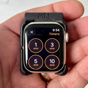 May include: A smartwatch encased in a black protective bumper frame. The screen displays a timer interface with four circular buttons labelled 1 MIN, 3 MIN, 5 MIN, and 10 MIN, along with the current time 9:34 and the text Timers at the top.
