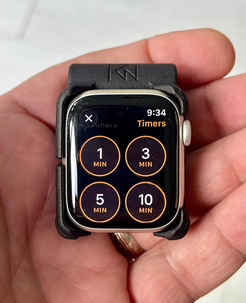 May include: A smartwatch encased in a black protective bumper frame. The screen displays a timer interface with four circular buttons labelled 1 MIN, 3 MIN, 5 MIN, and 10 MIN, along with the current time 9:34 and the text Timers at the top.