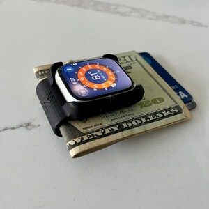 May include: A black silicone watch strap adapter securing a folded twenty-pound note and a blue debit card. The smartwatch screen features a circular weather interface displaying 81 degrees, the time 8:27, and a temperature range of 80 to 90 degrees.