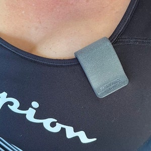 May include: A matte grey U-shaped clip attached to the neckline of a black athletic top. The clip features embossed text that reads PATENT PENDING and PIRANHA-MED.COM. The athletic top has a white logo visible on the front chest area.