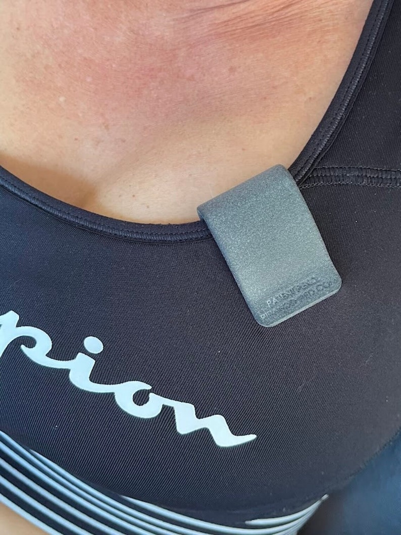 May include: A matte grey U-shaped clip attached to the neckline of a black athletic top. The clip features embossed text that reads PATENT PENDING and PIRANHA-MED.COM. The athletic top has a white logo visible on the front chest area.