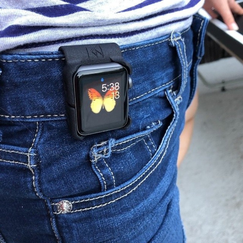 May include: A black silicone protective case holding a smartwatch attached to the waistband of dark blue denim jeans. The watch face displays a yellow and orange butterfly graphic, the time 5:38, and the number 13 in a digital font format.