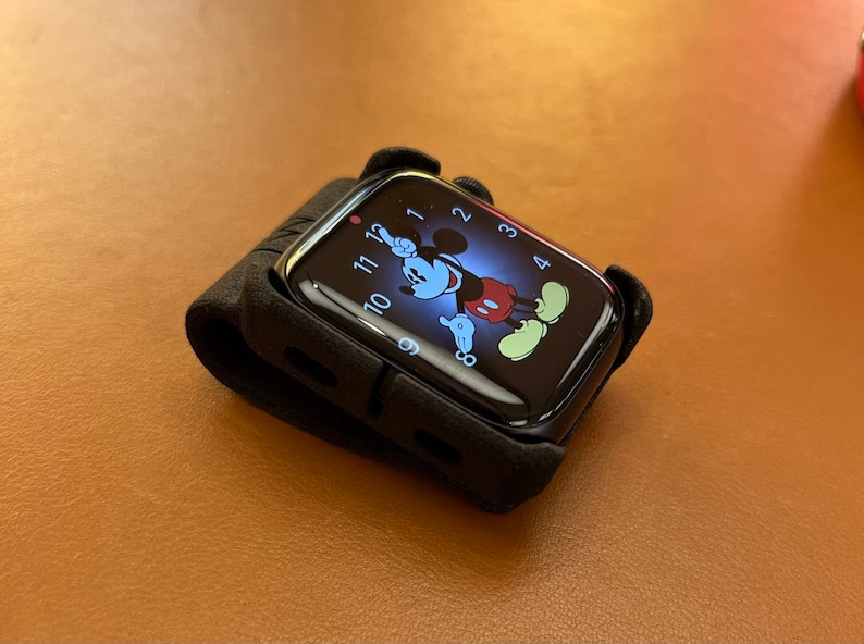 May include: A black smartwatch encased in a dark protective cover. The digital display features a graphic of Mickey Mouse with his arms serving as the clock hands, set against a dark blue background with white numerals from one to twelve.