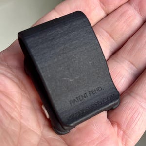 May include: A small, matte black U-shaped clip with a textured surface. The front face features the embossed text 'PATENT PEND.' and 'RIPANDSHRED.COM' near the bottom edge. A small white circular component is visible on the side of the clip.