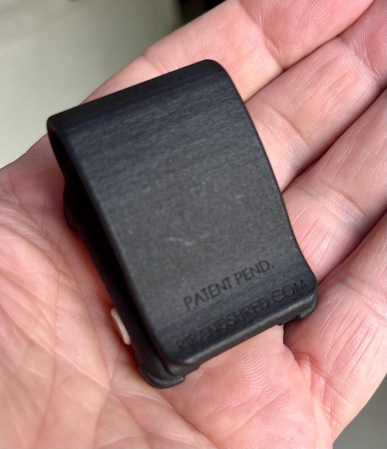 May include: A small, matte black U-shaped clip with a textured surface. The front face features the embossed text 'PATENT PEND.' and 'RIPANDSHRED.COM' near the bottom edge. A small white circular component is visible on the side of the clip.