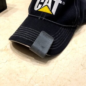 May include: A black baseball cap with white stitching and the CAT logo embroidered in white and yellow on the front. A dark grey clip is attached to the brim, featuring the text PATENT PEND. and the website address PIPANDSHRED.COM embossed on it.