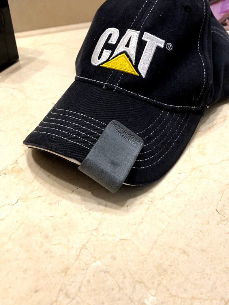 May include: A black baseball cap with white stitching and the CAT logo embroidered in white and yellow on the front. A dark grey clip is attached to the brim, featuring the text PATENT PEND. and the website address PIPANDSHRED.COM embossed on it.