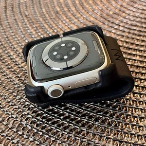 May include: A silver-toned smartwatch with a circular sensor array on the back, featuring a digital crown and a side button, secured within a black leather-like holder that has a small embossed logo on the side edge of the material.
