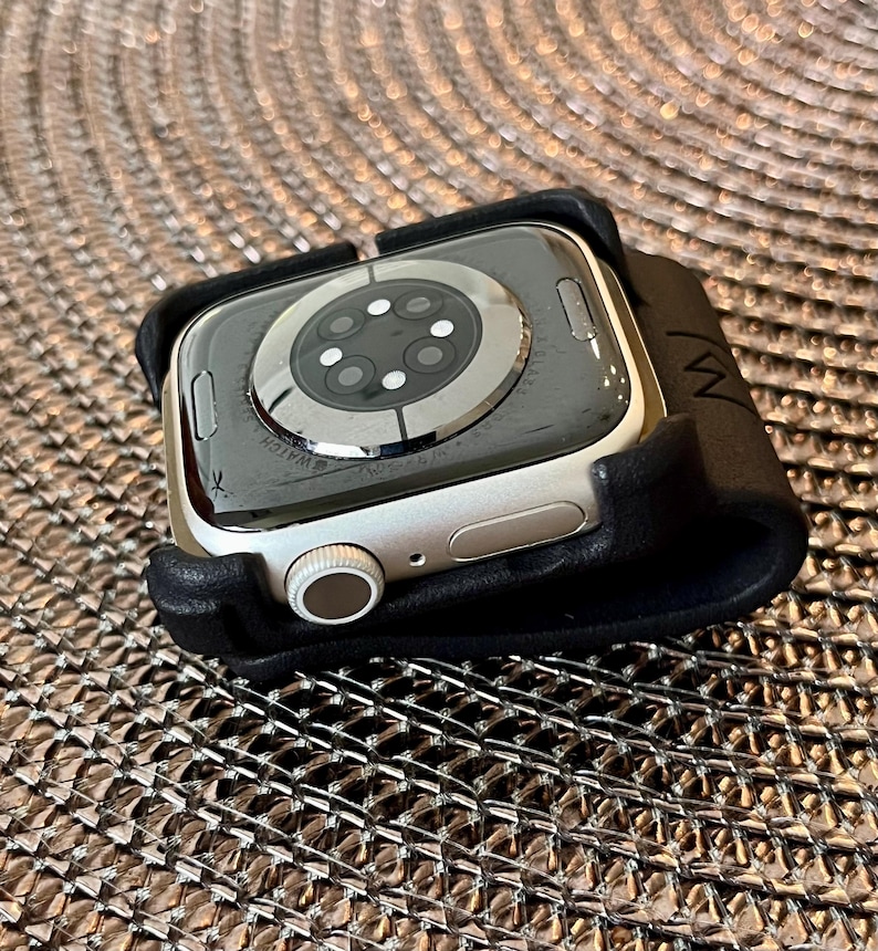 May include: A silver-toned smartwatch with a circular sensor array on the back, featuring a digital crown and a side button, secured within a black leather-like holder that has a small embossed logo on the side edge of the material.