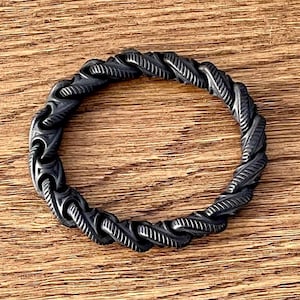 May include: A circular ring featuring a dark, oxidized finish with a continuous, twisted rope-like texture. The surface of the band displays deep grooves and raised diagonal ridges that create a repeating pattern around the entire circumference.