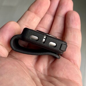 May include: A black silicone protective case for a small electronic device, featuring a curved loop design on one side and two metallic silver buttons on the side edge. The case has a matte, textured finish and a compact, rectangular shape.