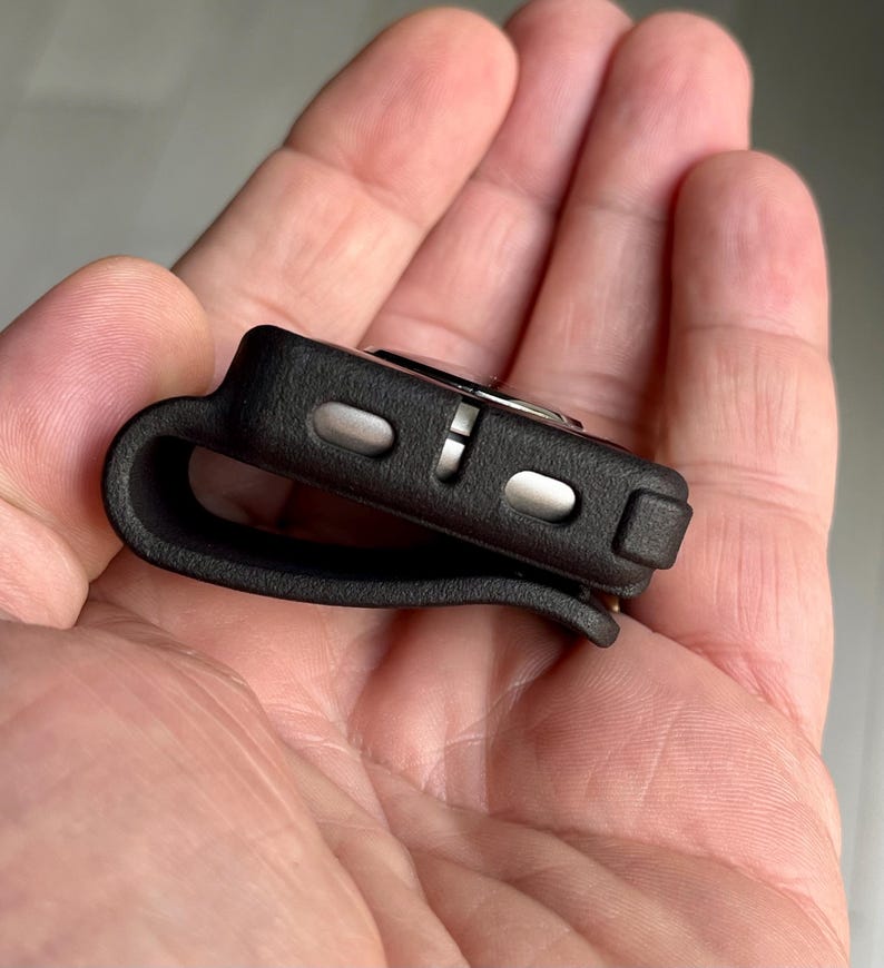May include: A black silicone protective case for a small electronic device, featuring a curved loop design on one side and two metallic silver buttons on the side edge. The case has a matte, textured finish and a compact, rectangular shape.
