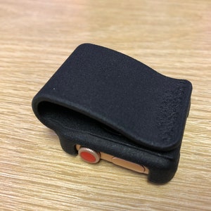 May include: A black matte protective cover for a smartwatch, featuring a textured finish and a flexible clip design. The cover is fitted over a gold-toned watch case that displays a prominent red circular digital crown on the side.