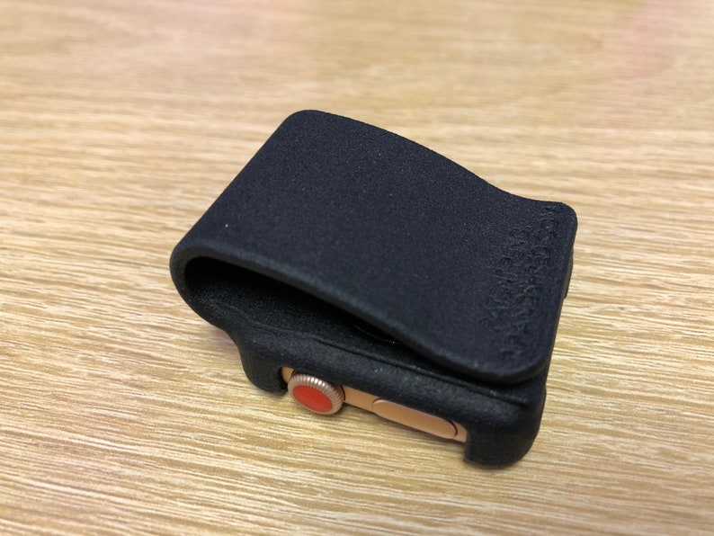 May include: A black matte protective cover for a smartwatch, featuring a textured finish and a flexible clip design. The cover is fitted over a gold-toned watch case that displays a prominent red circular digital crown on the side.