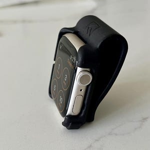 May include: A silver smartwatch with a black digital screen displaying timer options is held in a black, curved, matte-textured stand. The stand features a small embossed logo on the top edge and holds the watch at an upright angle.