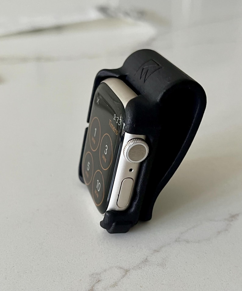 May include: A silver smartwatch with a black digital screen displaying timer options is held in a black, curved, matte-textured stand. The stand features a small embossed logo on the top edge and holds the watch at an upright angle.