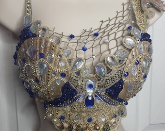 May include: A gold and navy blue embellished top with a seashell design. The top is adorned with iridescent and blue gemstones, pearls, and gold trim. A decorative starfish is attached to the shoulder. The top is displayed on a mannequin.