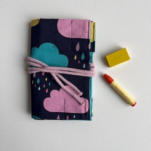 Könnte beinhalten: Eine dunkelblaue Stofftasche mit einem Wolken- und Regentropfenmuster. Die Tasche ist mit einer rosa Schnur gebunden und enthält einen gelben Radiergummi und einen roten Wachsmalstift mit dem Text "WACHSMALSTIFT" darauf.