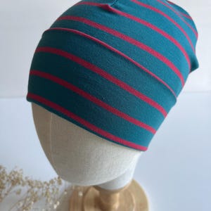 Op de afbeelding: Een muts met een blauwgroene basiskleur en horizontale rode strepen. De muts is gemaakt van een zachte, flexibele stof en wordt getoond op een etalagepophoofd om de ronde vorm en de pasvorm goed weer te geven.