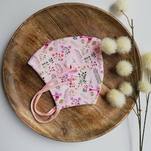 Può includere: Una maschera facciale rosa con stampa floreale e di uccelli. La maschera ha lacci rosa ed è appoggiata su un piatto di legno.