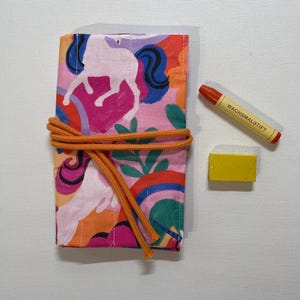 Op de afbeelding: Een rechthoekige stoffen oproletui met een kleurrijk patroon van eenhoorns, regenbogen en bladeren, vastgezet met een oranje koord. Naast de etui liggen een oranje waskrijtje met het opschrift WACHSMALSTIFT en een kleine gele gum.