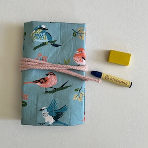 Op de afbeelding: Een blauwe stoffen tas met een vogelpatroon, vastgebonden met een roze koord. Een gele gum en een blauwe stift met de tekst "STOCKMAR" liggen op een wit oppervlak.