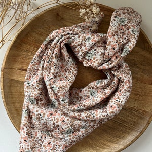 Puede incluir: Un pañuelo de tela blanco con un estampado floral en tonos rosa, marrón y verde. El pañuelo está doblado y se muestra sobre una superficie de madera.