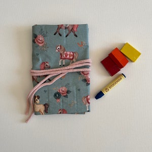 Op de afbeelding: Een blauwe stoffen tas met een paardenprint en een roze trekkoord. De tas is gevuld met kleurpotloden en een blauw kleurpotlood ligt ernaast.