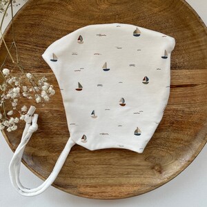 Könnte beinhalten: Ein weißer Baby-Mützchen mit einer Binde zum Verschließen, mit einem Muster aus kleinen Segelbooten und Wellen in Blautönen, Braun und Beige.