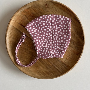 Puede incluir: Una mascarilla facial de tela rosa con un patrón de lunares blancos. La mascarilla tiene lazos para atar detrás de la cabeza.