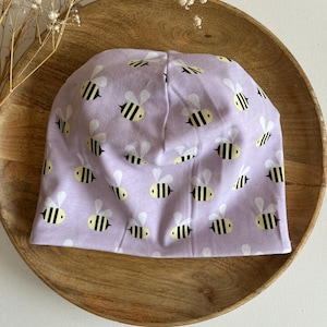 Op de afbeelding: Een lavendelkleurige beanie-muts met een herhaald patroon van gele en zwarte bijen. De muts is gemaakt van zachte stof en is ontworpen om goed op het hoofd te passen. De muts wordt gepresenteerd op een houten bord.