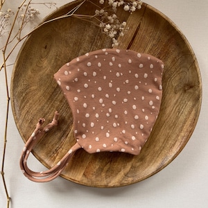 Peut inclure: Un bonnet en tissu marron à pois blancs et deux longues attaches. Le bonnet est posé sur une assiette en bois.