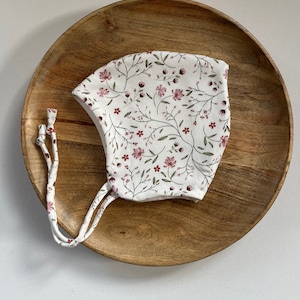 Può includere: Un berretto bianco con un motivo floreale in tonalità rosa e verde. Il berretto ha due lunghi lacci che possono essere legati sotto il mento.