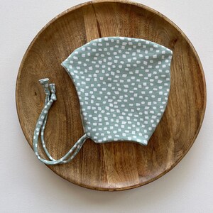 Pode incluir: Um gorro de bebê verde claro com um padrão de bolinhas brancas. O gorro tem laços e é apresentado em um prato de madeira. O gorro é feito de um tecido macio.