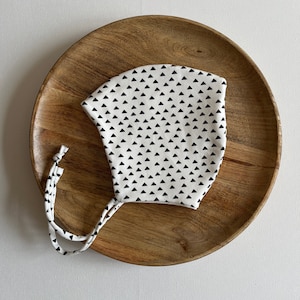 Pode incluir: Um gorro de bebé branco com um padrão de triângulos pretos. O gorro tem laços que podem ser amarrados sob o queixo.