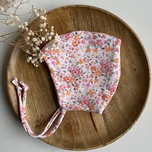 Op de afbeelding: Een bloemen baby muts met een witte achtergrond en een patroon van roze, oranje en gele bloemen. De muts heeft strikkoorden en wordt gepresenteerd op een houten bord.