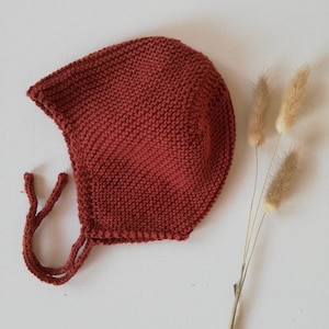 Puede incluir: Un gorro de bebé tejido de color óxido con lazos. El gorro está hecho de una tela de punto texturizada y tiene una forma redondeada con un borde puntiagudo. Hierba ornamental seca a la derecha del gorro.