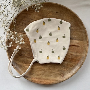 Pode incluir: Um gorro de bebé branco com fecho de nó. O gorro tem um padrão de maçãs verdes e peras amarelas.