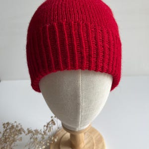 Könnte beinhalten: Eine leuchtend rote Strickmütze. Die Mütze wird auf einem Schaufensterkopf präsentiert, der ihr geripptes Design und ihre enge Passform hervorhebt. Die Mütze besteht aus einem weichen, warmen Material, ideal für kaltes Wetter.