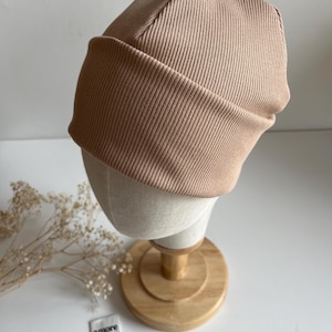 Könnte beinhalten: Eine beige gerippte Strickmütze mit umgeschlagenem Rand. Die Mütze wird auf einem Schaufensterkopf präsentiert. Zwei kleine Etiketten sind sichtbar, eines mit den Worten "amore mio" und das andere mit dem Wort "hello" und einem Herzsymbol.