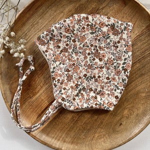 Op de afbeelding: Een witte muts met een bloemenprint in bruine en roze tinten. De muts heeft een strik die onder de kin kan worden geknoopt.