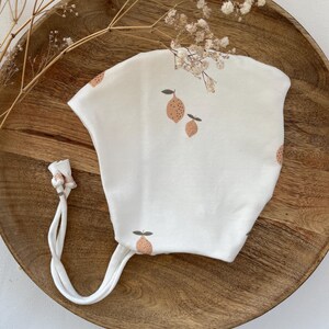 Puede incluir: Un gorro blanco para bebé con estampado de limón. El gorro tiene un lazo en la parte posterior.