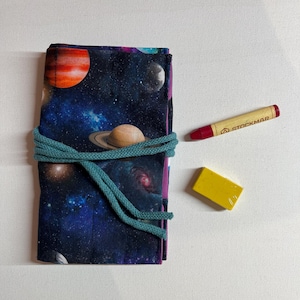 Pode incluir: Um estojo de lápis de tecido dobrado com um padrão de tema espacial, amarrado com um cordão azul-petróleo. Um giz de cera vermelho com "STOCKMAR" impresso e um bloco de cera amarelo estão ao lado do estojo. O estojo apresenta planetas e galáxias.