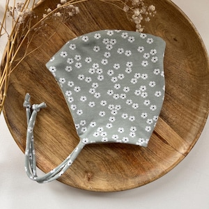 Op de afbeelding: Een saliegroene baby muts met een wit bloemenpatroon. De muts heeft twee lange banden.