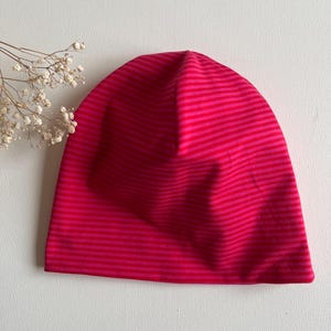 Puede incluir: Un gorro de rayas rosas y rojas. El gorro está hecho de un material suave y presenta rayas horizontales en diferentes tonos de rosa y rojo. El gorro es de un estilo sencillo y ajustado, adecuado para el clima frío.